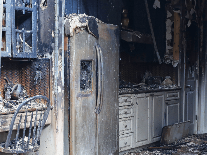 Kitchen severely damaged by fire, with charred cabinets, a melted refrigerator, and ash-covered countertops and flooring.