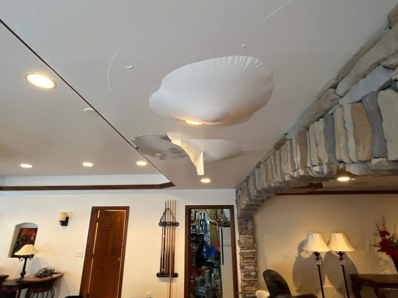 Living room ceiling with severe water damage causing large, balloon-like bulges in the drywall, surrounded by recessed lighting and a stone archway.