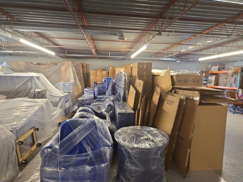 Warehouse interior with stacks of furniture, cardboard boxes, and equipment covered in protective plastic wrap.